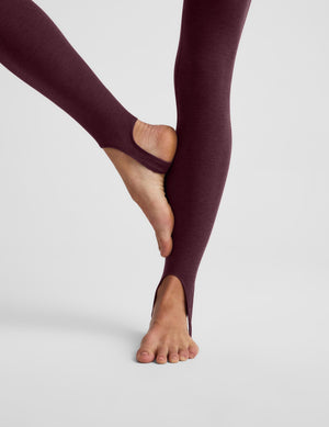 A close-up of a person's legs wearing dark plum-colored split-heel tights against a light gray studio background.