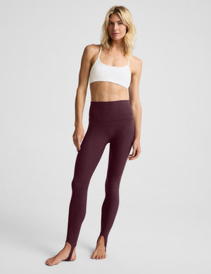 A woman modeling high-waisted leggings in a deep plum purple color, paired with a white sports bra, against a light gray studio background.