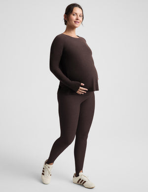 A pregnant model wearing a matching dark brown knit long-sleeve top and leggings set, paired with white sneakers, standing against a light gray studio background.