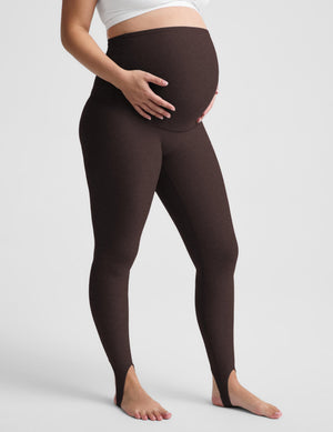 A pregnant model wearing dark brown maternity leggings standing barefoot against a light gray studio background.