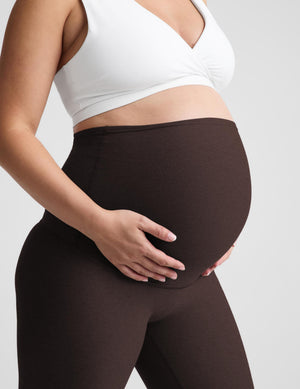 Pregnant model wearing a white sports bra and dark brown high-waisted maternity leggings, holding her baby bump against a light gray background.