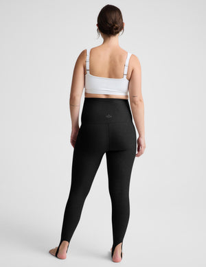 A pregnant model wearing black maternity leggings and a white sports bra, standing against a light gray studio background.