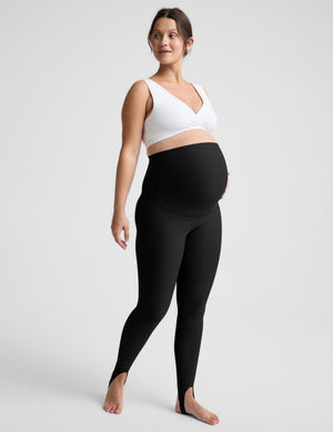 A pregnant model wearing black maternity leggings and a white sports bra, standing against a light gray studio background.