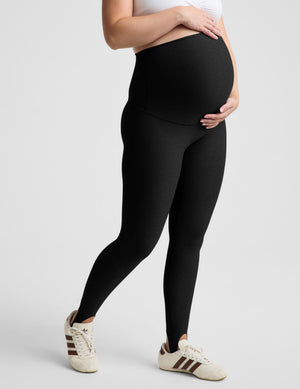 A pregnant model wearing black maternity leggings and a white sports bra, standing against a light gray studio background.
