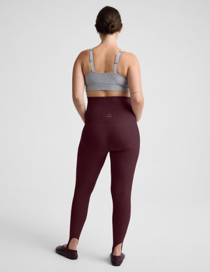 A woman viewed from the back wearing high-waisted burgundy leggings and a light gray sports bra against a light gray studio background.