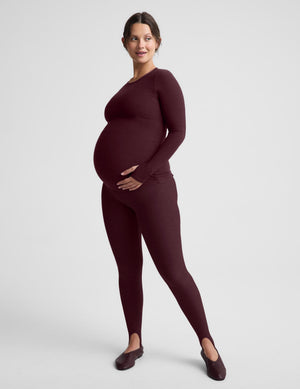 A pregnant model wearing a matching long-sleeve maternity top and leggings set in a deep burgundy color, standing against a light gray studio background.