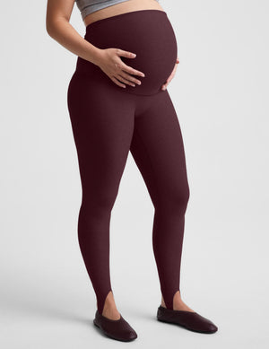 A pregnant model wearing high-waisted, maternity leggings in a deep burgundy color, standing against a light gray studio background.