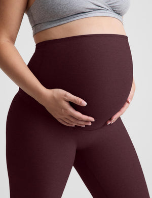 Pregnant model wearing high-waisted burgundy maternity leggings and a gray sports bra, holding her baby bump against a light gray background.