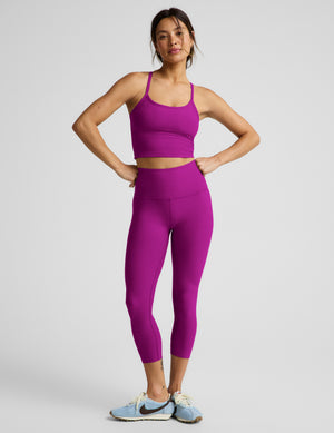 Woman wearing a purple cropped tank top and purple capri leggings against a plain background. 