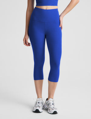 A woman modeling a matching royal blue activewear set consisting of a sports bra and high-waisted capri leggings, paired with white and gray sneakers, against a light gray studio background.