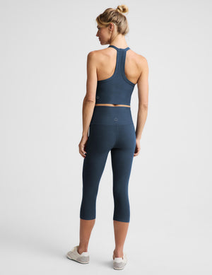 A woman is shown from behind wearing a matching dark blue athletic set consisting of a racerback crop top and high-waisted capri leggings, paired with white sneakers, against a light gray studio background.