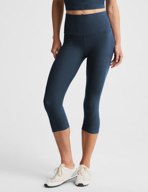 A model wearing high-waisted, dark blue-gray capri leggings and white sneakers, shown from the waist down against a light gray studio background.