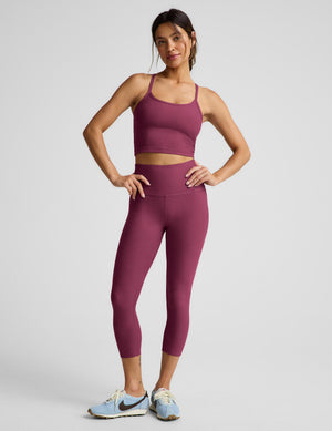 A woman modeling a matching red activewear set consisting of a scoop-neck crop top and high-waisted capri leggings, paired with light blue running shoes, against a light gray studio background.
