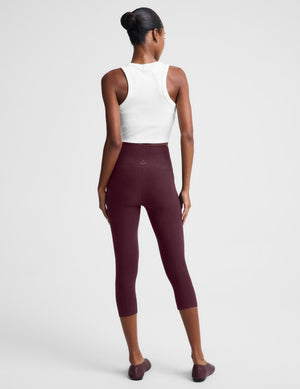 A woman modeling a matching activewear set consisting of a white sleeveless crop top and high-waisted burgundy capri leggings, paired with burgundy flats, against a light gray studio background.
