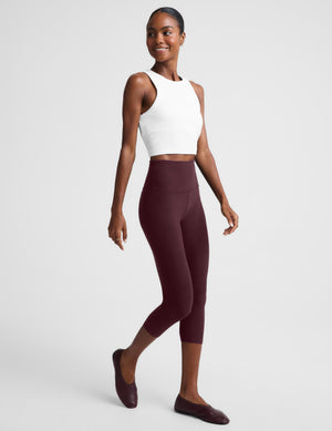 A woman modeling a matching activewear set consisting of a white sleeveless crop top and high-waisted burgundy capri leggings, paired with burgundy flats, against a light gray studio background.