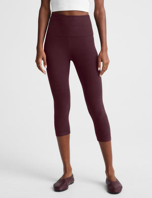 A woman modeling a matching activewear set consisting of a white sleeveless crop top and high-waisted burgundy capri leggings, paired with burgundy flats, against a light gray studio background.