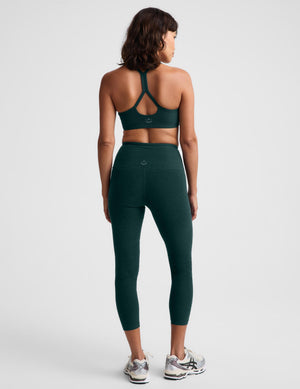 Woman wearing a green racerback sports bra and matching high waisted capri leggings against a plain studio background. 