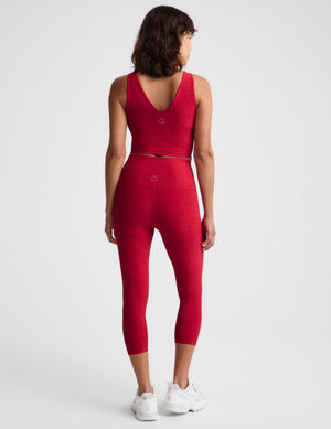 A woman viewed from the back wearing a matching red activewear set consisting of a sleeveless crop top and high-waisted capri leggings, paired with white sneakers, against a light gray studio background.