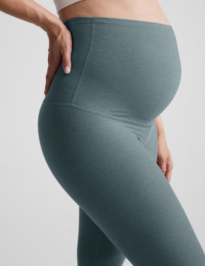 Woman wearing a white bra and green maternity capri leggings against a plain background. 