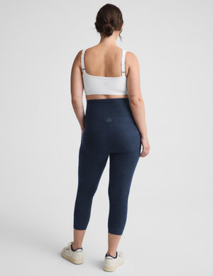 A woman is shown from behind wearing a white sports bra and navy blue high-waisted leggings, standing against a light gray studio background.