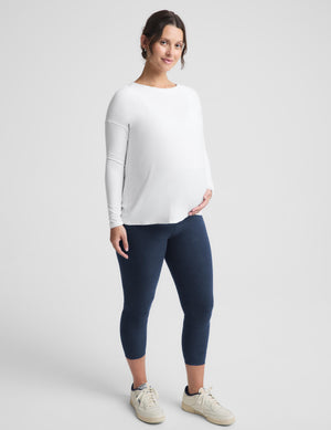 A pregnant model wearing a white long-sleeve maternity top, navy blue maternity leggings, and off-white sneakers, standing against a light gray studio background.