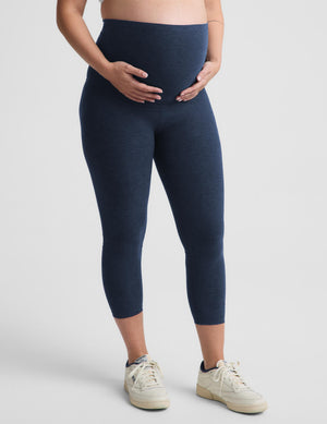 A pregnant model wearing navy blue maternity leggings and off-white sneakers, standing against a light gray studio background.