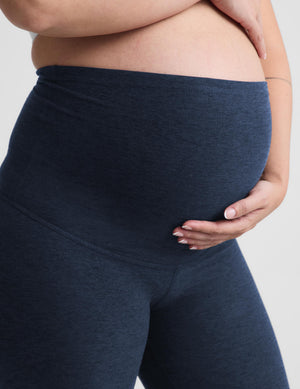 Close-up of a pregnant model wearing dark heather navy maternity leggings, holding her baby bump with one hand against a light gray background.