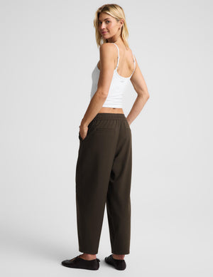 A woman modeling a pair of dark brown wide-leg cropped pants with an elastic waistband, paired with a white tank top and black flats, shown from a three-quarters back view against a light gray studio background.