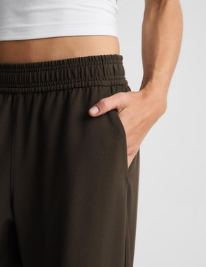 Close-up of a person wearing dark brown wide-leg sweatpants with an elastic waistband and side pocket, with one hand in the pocket. A white cropped tank top is partially visible at the top against a light gray background.