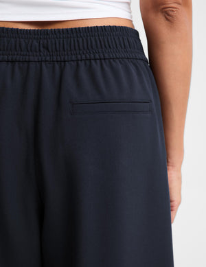 Close-up of a person wearing dark navy wide-leg sweatpants with an elastic waistband and a side pocket, paired with a white cropped top against a light gray background.