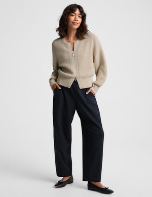 A woman modeling a light beige ribbed zip-front cardigan with balloon sleeves, paired with dark wide-leg trousers and black flats, against a light gray studio background.