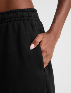Close-up of a person wearing black sweatpants, showing the side pocket and hand with white manicured nails resting in the pocket against a light gray background.
