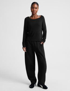 A model wearing a black long-sleeve top and matching black wide-leg sweatpants, standing against a light gray studio background.