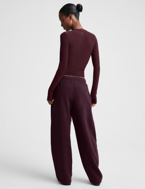 A model wearing a matching dark burgundy two-piece lounge set consisting of a long-sleeve top and wide-leg sweatpants, photographed against a neutral light gray studio background.