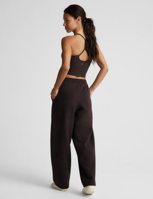 A woman modeling a dark brown activewear set consisting of a crop tank and matching barrel leg sweatpants, paired with white sneakers, against a light gray studio background.