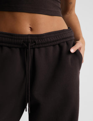 A woman modeling a dark brown activewear set consisting of a crop tank and matching barrel leg sweatpants, paired with white sneakers, against a light gray studio background.