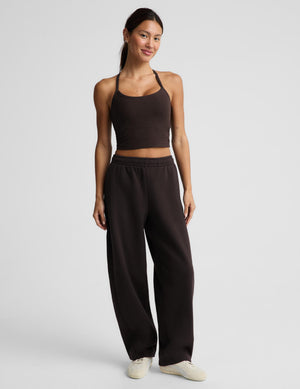 A woman modeling a dark brown activewear set consisting of a crop tank and matching barrel leg sweatpants, paired with white sneakers, against a light gray studio background.