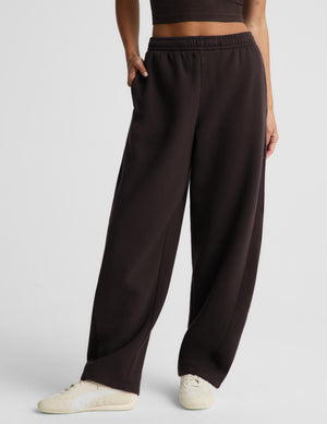 A woman modeling a dark brown activewear set consisting of a crop tank and matching barrel leg sweatpants, paired with white sneakers, against a light gray studio background.