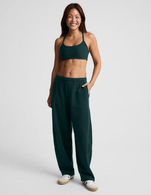 A woman modeling a matching dark green activewear set consisting of a sports bra and wide-leg sweatpants, paired with white sneakers, against a light gray studio background.