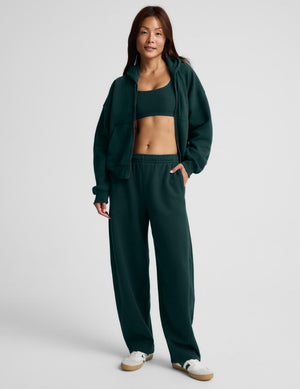 A woman modeling a dark green athletic tracksuit consisting of a cropped zip-up hoodie, a matching sports bra, and wide-leg sweatpants, paired with white sneakers, against a light gray studio background.