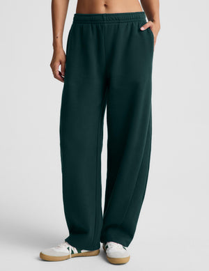 A person wearing dark green wide-leg sweatpants with an elastic waistband and side pockets, paired with white sneakers, shown from the waist down against a light gray studio background.