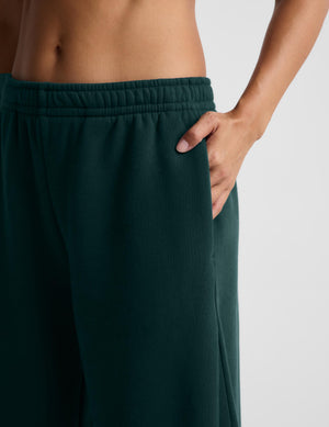 Close-up of a person wearing dark forest green sweatpants, showing the side pocket with a hand in the pocket and the elastic waistband.