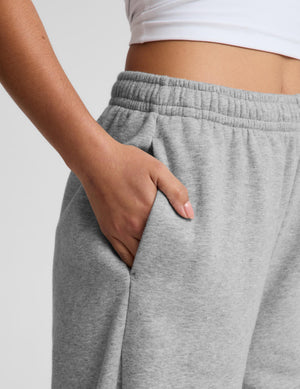 Close-up of a person wearing light heather gray sweatpants with an elastic waistband and side pocket, with one hand in the pocket and a white cropped top visible at the top.