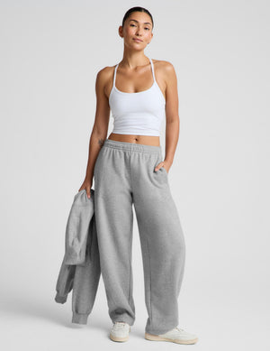 A woman modeling a pair of light gray wide-leg sweatpants with a white ribbed tank top and white sneakers, holding a matching gray sweatshirt against a light gray studio background.