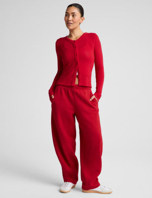 A woman modeling a matching red knit lounge set consisting of a cropped long-sleeve top with a button-front detail and relaxed wide-leg pants, paired with white sneakers, against a light gray studio background.