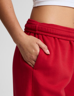 Close-up of a person wearing bright red sweatpants with a ribbed elastic waistband and a side pocket, with one hand in the pocket. A white cropped tank top is partially visible at the top against a light gray background.