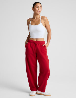 A woman modeling a pair of loose-fit red sweatpants with a white ribbed camisole top and white sneakers, standing against a light gray studio background.