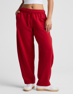 A person wearing loose red sweatpants with a drawstring waist, paired with white sneakers, shown from the waist down against a light gray studio background.
