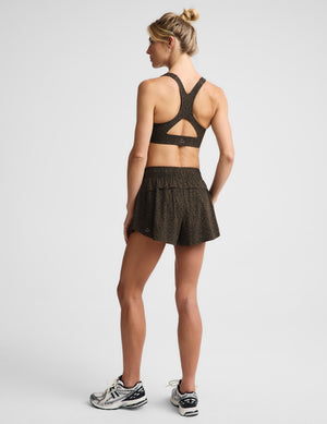 A woman modeling a matching dark brown and black speckled activewear set, consisting of a sports bra and shorts, paired with gray running shoes, against a light gray studio background.