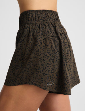 A close-up of a model wearing a dark olive green athletic skirt with a black leopard print pattern and a small white embroidered logo near the hem.